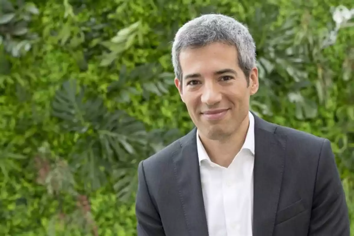 Un hombre con cabello canoso y traje oscuro sonríe frente a un fondo de vegetación verde. Un hombre con cabello canoso y traje oscuro sonríe frente a un fondo de vegetación verde.