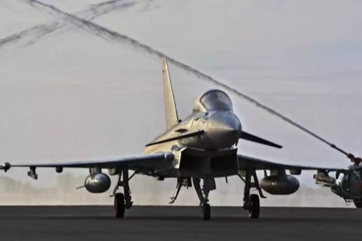Un avión de combate en una pista de aterrizaje siendo rociado con agua desde un vehículo cercano. Un avión de combate en una pista de aterrizaje siendo rociado con agua desde un vehículo cercano.