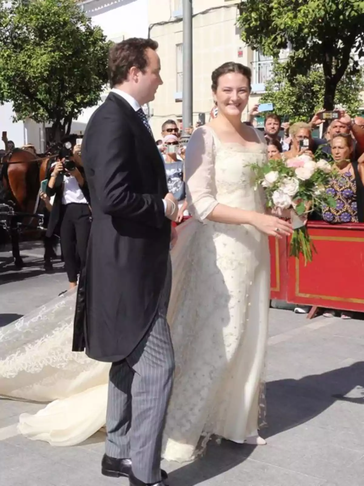 Pareja vestida de novios caminando al aire libre mientras la novia sostiene un ramo de flores y varias personas los observan y toman fotos Pareja vestida de novios caminando al aire libre mientras la novia sostiene un ramo de flores y varias personas los observan y toman fotos