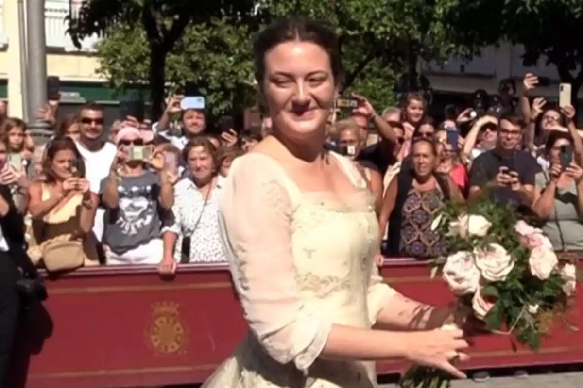 Mujer con vestido claro y ramo de flores sonríe mientras camina frente a una multitud que la fotografía en un evento al aire libre Mujer con vestido claro y ramo de flores sonríe mientras camina frente a una multitud que la fotografía en un evento al aire libre