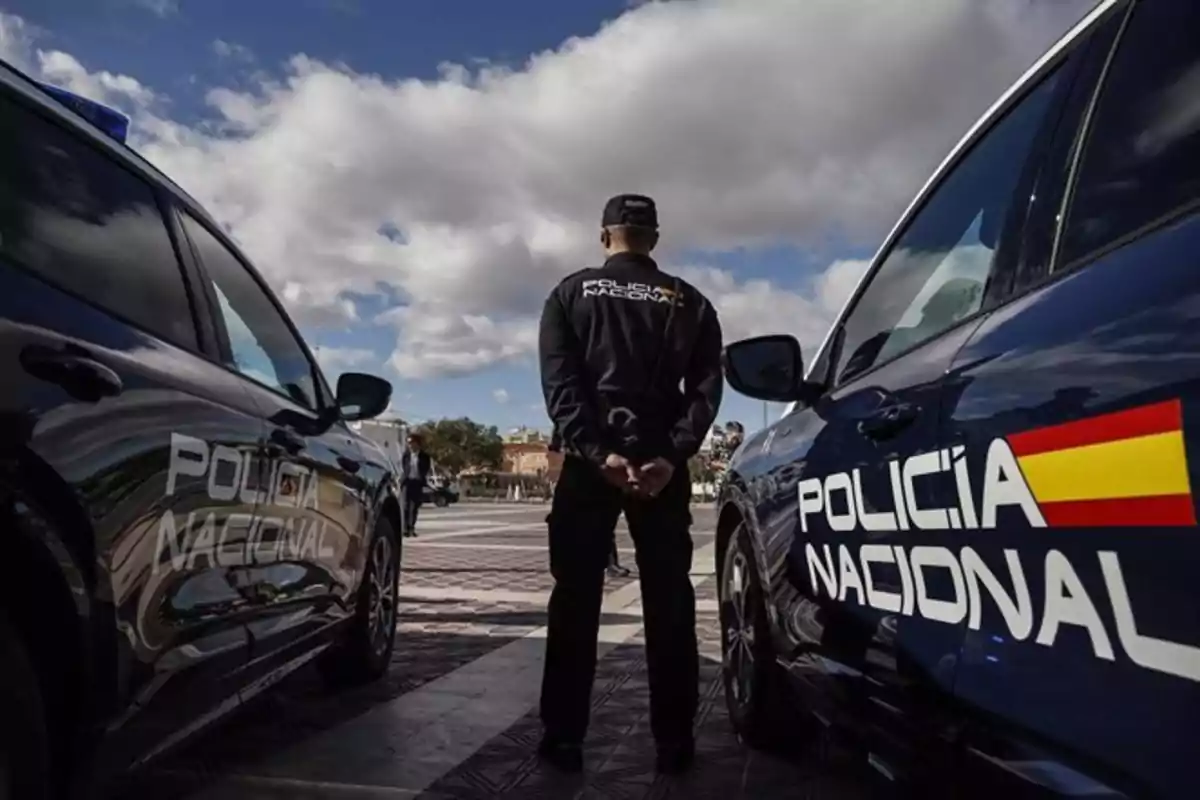 Un agente de policía de pie entre dos coches patrulla de la Policía Nacional en un día soleado con nubes en el cielo.