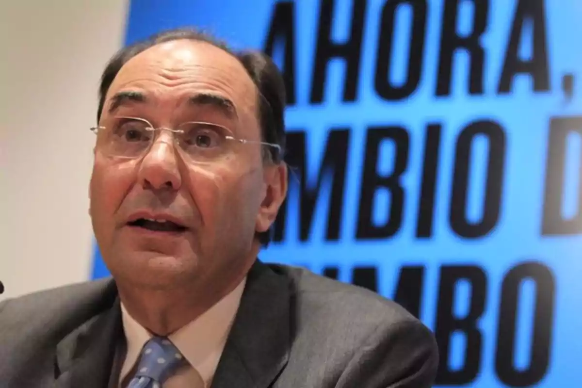 Un hombre con gafas y traje habla frente a un cartel azul con texto en letras grandes. Un hombre con gafas y traje habla frente a un cartel azul con texto en letras grandes.