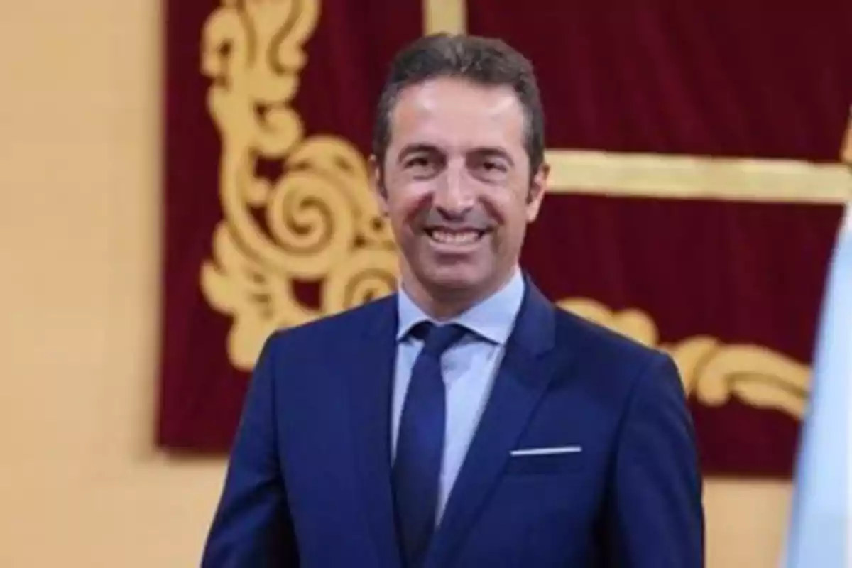 Hombre de cabello corto y oscuro con traje azul y corbata posando y sonriendo frente a un fondo decorativo rojo y dorado Hombre de cabello corto y oscuro con traje azul y corbata posando y sonriendo frente a un fondo decorativo rojo y dorado