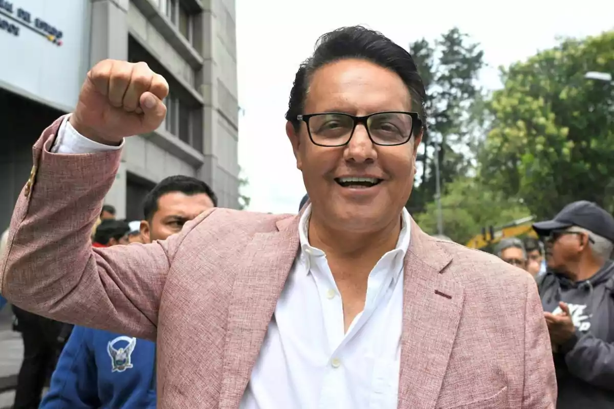 Hombre con gafas y saco rosa sonríe y levanta el puño en señal de celebración mientras está al aire libre rodeado de personas Hombre con gafas y saco rosa sonríe y levanta el puño en señal de celebración mientras está al aire libre rodeado de personas