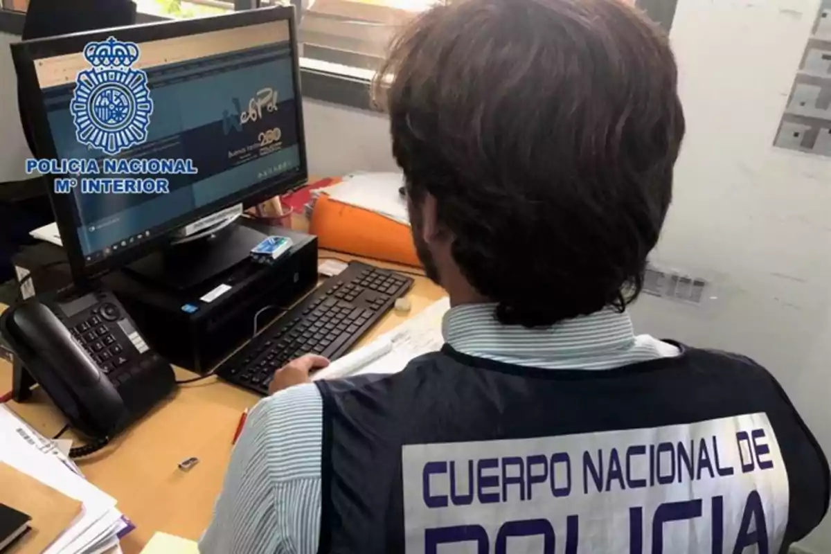Un agente del Cuerpo Nacional de Policía trabaja en un escritorio frente a una computadora, con un chaleco que lleva el nombre de la institución. Un agente del Cuerpo Nacional de Policía trabaja en un escritorio frente a una computadora, con un chaleco que lleva el nombre de la institución.