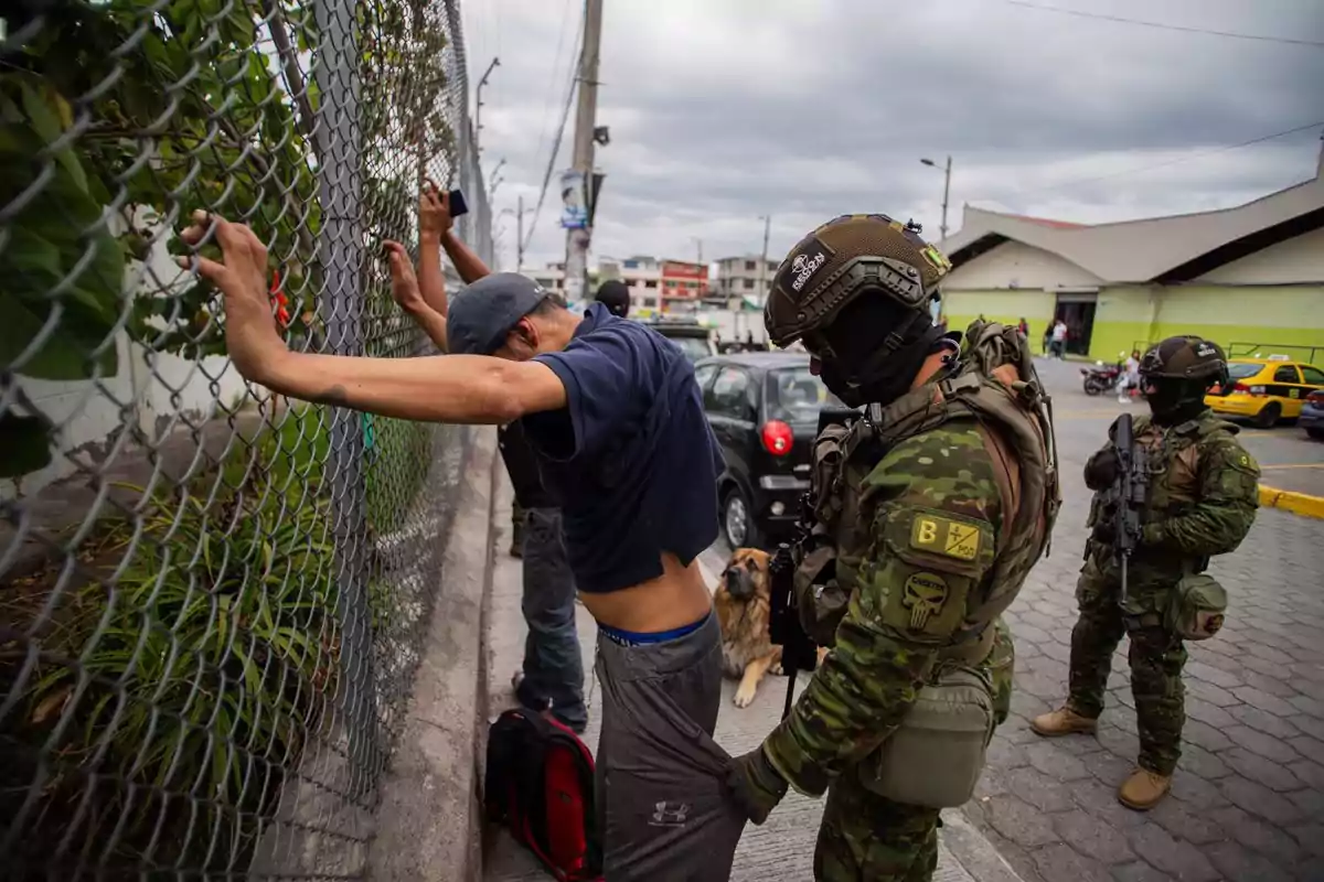 Un oficial militar revisa a un hombre apoyado en una cerca mientras otro oficial observa y un perro está cerca en una calle urbana.