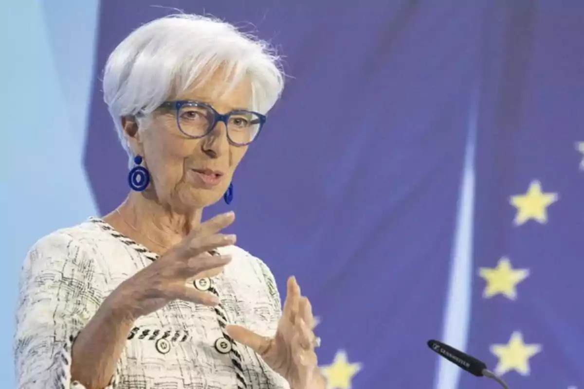 Una mujer de cabello blanco y gafas azules gesticula mientras habla frente a un fondo con estrellas amarillas.