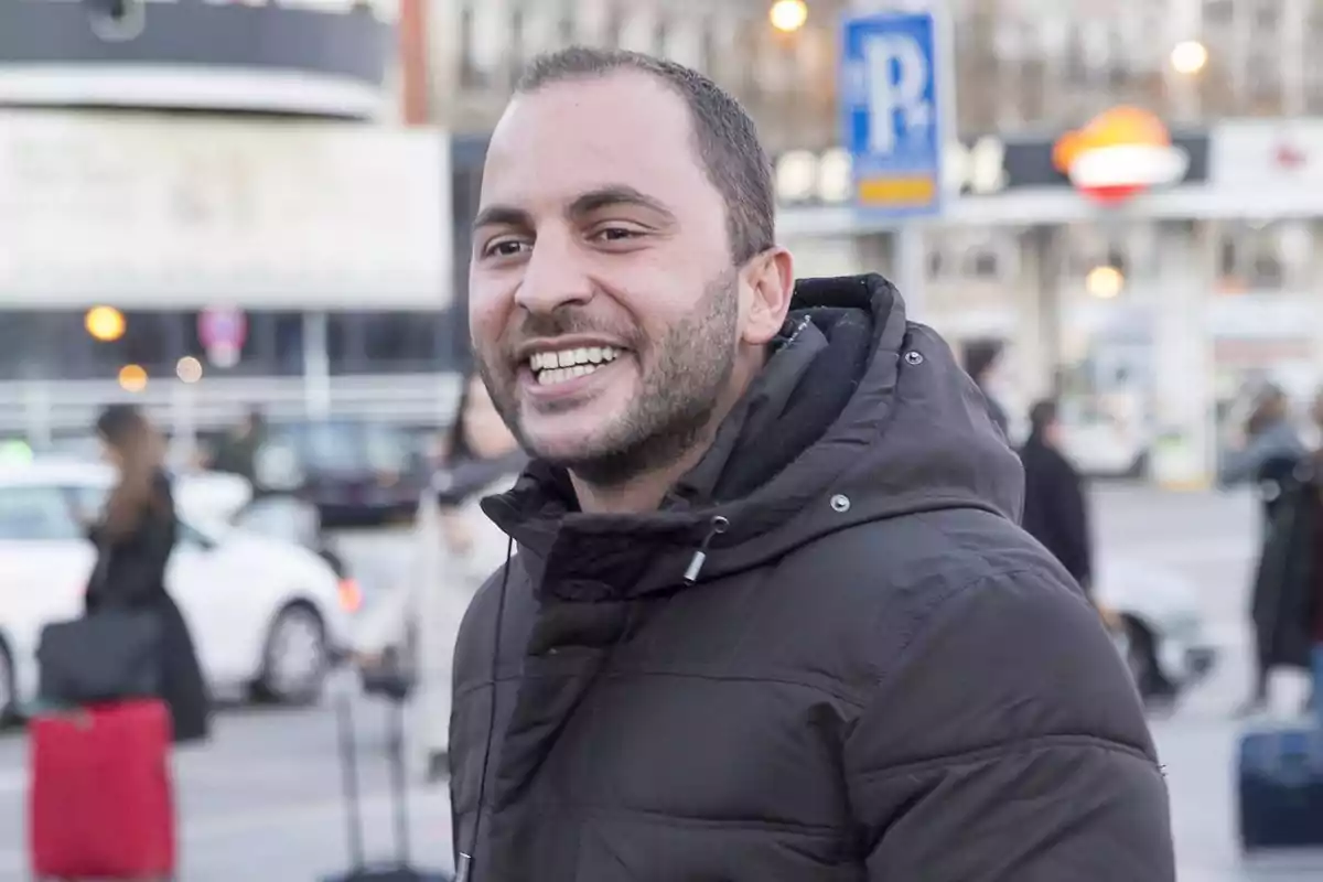 Un hombre sonriente con chaqueta oscura en un entorno urbano con personas y coches de fondo. Un hombre sonriente con chaqueta oscura en un entorno urbano con personas y coches de fondo.