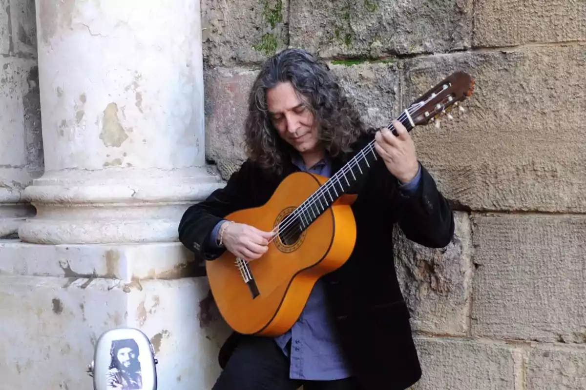 Un hombre con cabello largo y rizado toca una guitarra clásica mientras se apoya en una columna de piedra, con una funda de guitarra con una imagen en el suelo.