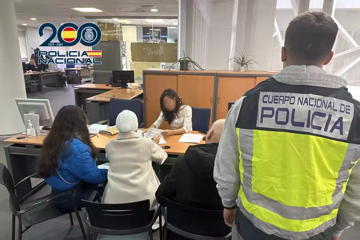 Un agente de policía con chaleco del Cuerpo Nacional de Policía observa a tres personas sentadas en una oficina mientras una mujer detrás de un escritorio revisa documentos.