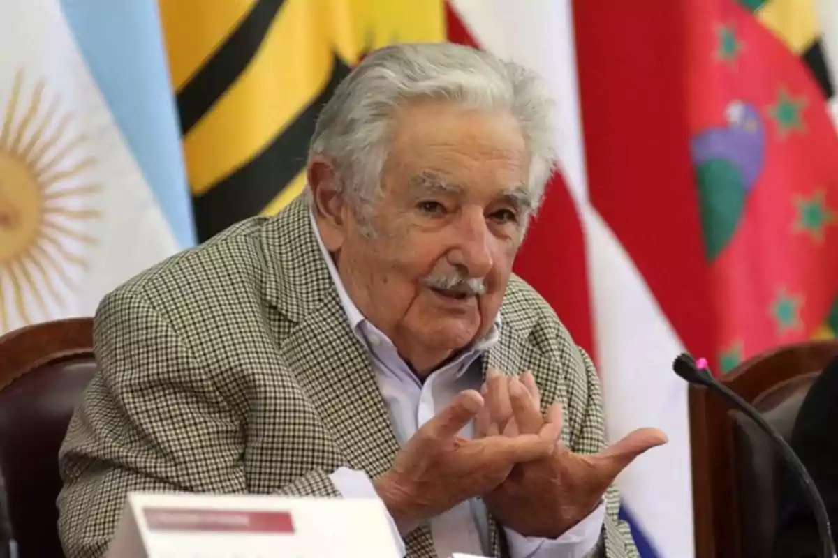Un hombre mayor con cabello canoso y bigote está sentado en una mesa, gesticulando con las manos, con varias banderas de fondo. Un hombre mayor con cabello canoso y bigote está sentado en una mesa, gesticulando con las manos, con varias banderas de fondo.