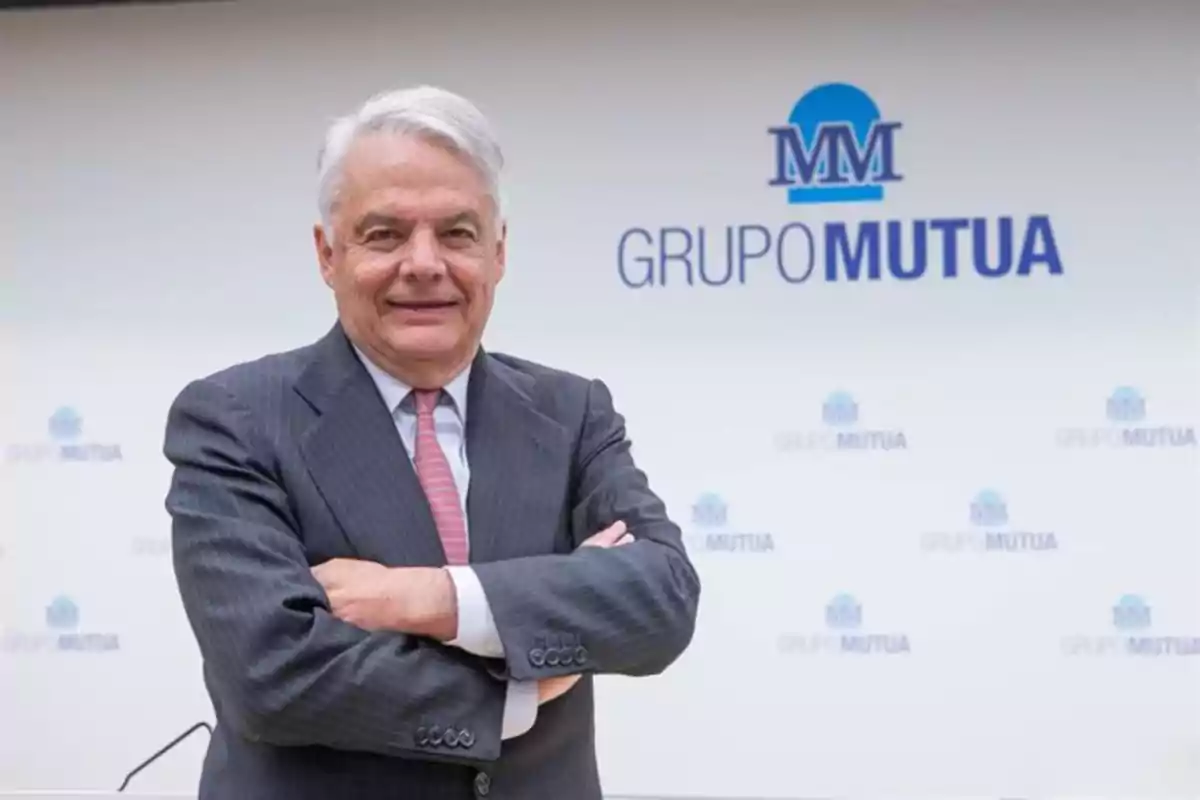 Hombre de cabello canoso con traje y corbata posando con los brazos cruzados frente a un fondo con el logo de Grupo Mutua
