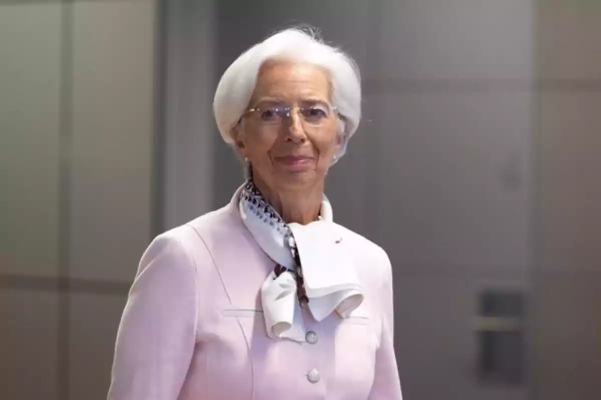 Mujer de cabello blanco y gafas con traje rosa claro y pañuelo al cuello en un entorno interior.