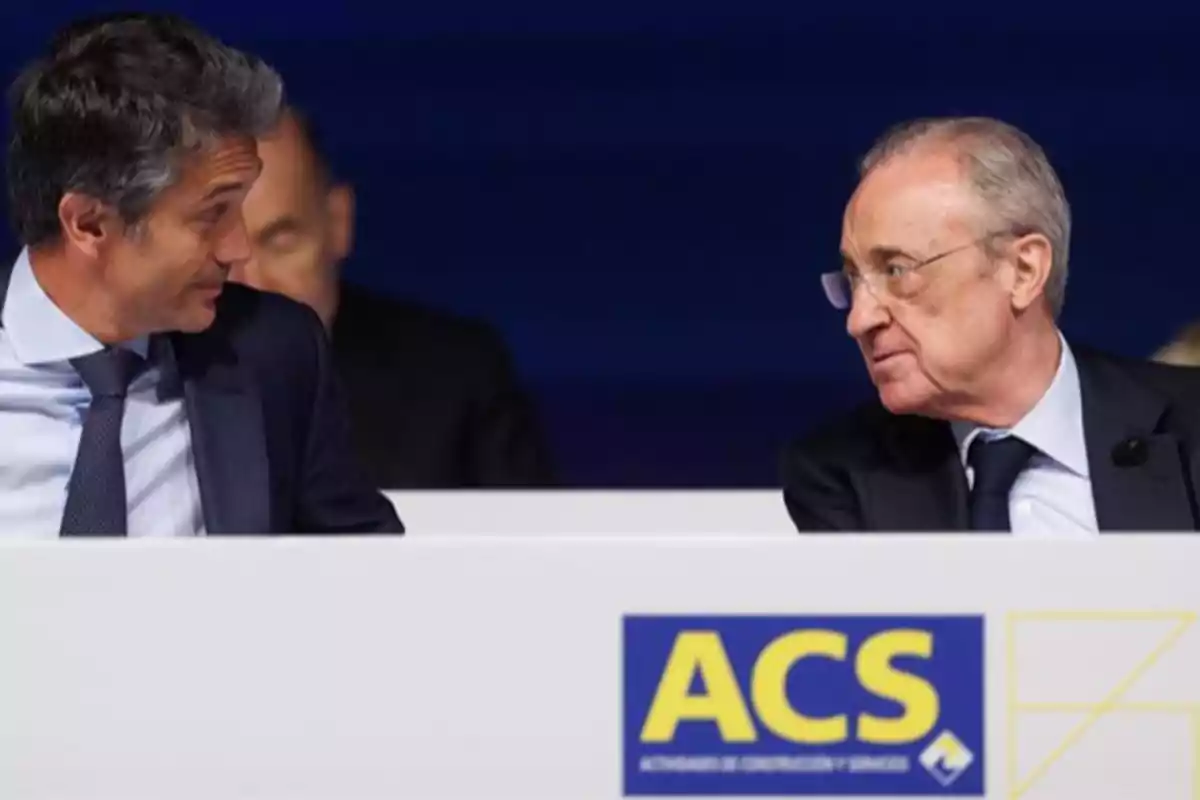 Dos hombres con traje conversan sentados detrás de un panel con el logo de ACS Dos hombres con traje conversan sentados detrás de un panel con el logo de ACS