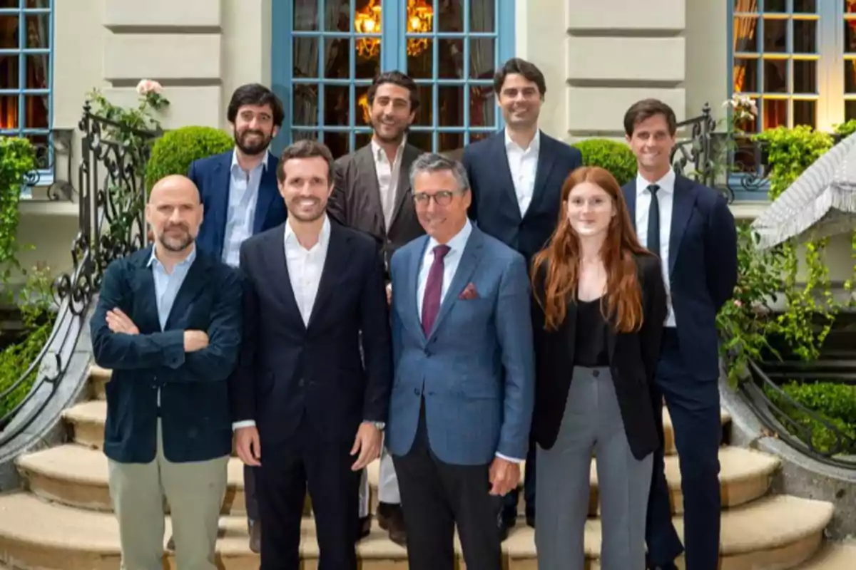 Un grupo de personas vestidas formalmente posando en una escalera frente a un edificio elegante con ventanas grandes y plantas decorativas.