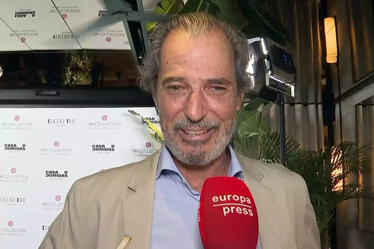 Hombre de cabello canoso y barba sonríe mientras sostiene un micrófono rojo de europa press en un evento con fondo de logotipos y plantas. Hombre de cabello canoso y barba sonríe mientras sostiene un micrófono rojo de europa press en un evento con fondo de logotipos y plantas.