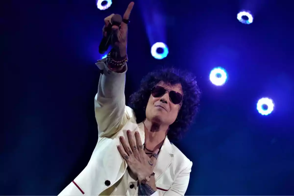 Un cantante con chaqueta blanca y gafas de sol en el escenario con luces azules de fondo.