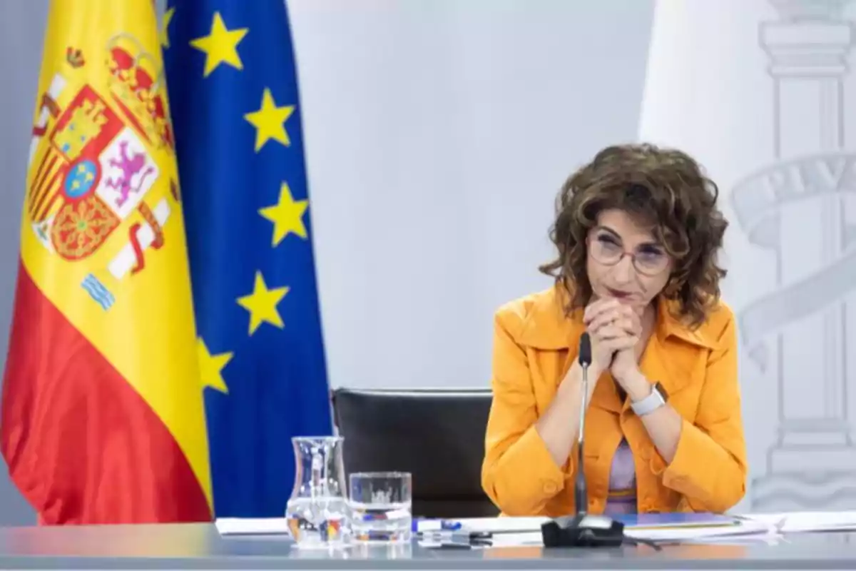 Mujer con gafas y chaqueta naranja sentada en una mesa frente a micrófono con banderas de España y la Unión Europea al fondo Mujer con gafas y chaqueta naranja sentada en una mesa frente a micrófono con banderas de España y la Unión Europea al fondo