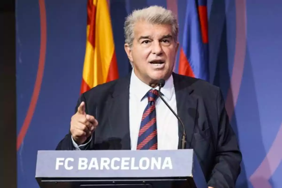 Hombre de traje hablando en un podio con el logo de FC Barcelona y banderas de fondo.