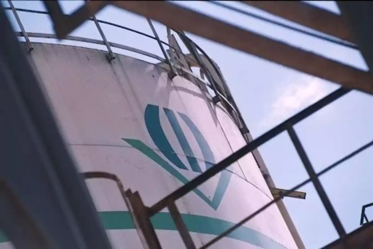 Tanque industrial con un logotipo azul y verde visto desde un ángulo bajo con estructuras metálicas en primer plano. Tanque industrial con un logotipo azul y verde visto desde un ángulo bajo con estructuras metálicas en primer plano.