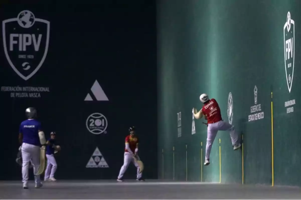 Jugadores de pelota vasca en acción dentro de un frontón, uno de ellos salta para golpear la pelota mientras otros observan, con uniformes de los equipos de Francia y España. Jugadores de pelota vasca en acción dentro de un frontón, uno de ellos salta para golpear la pelota mientras otros observan, con uniformes de los equipos de Francia y España.