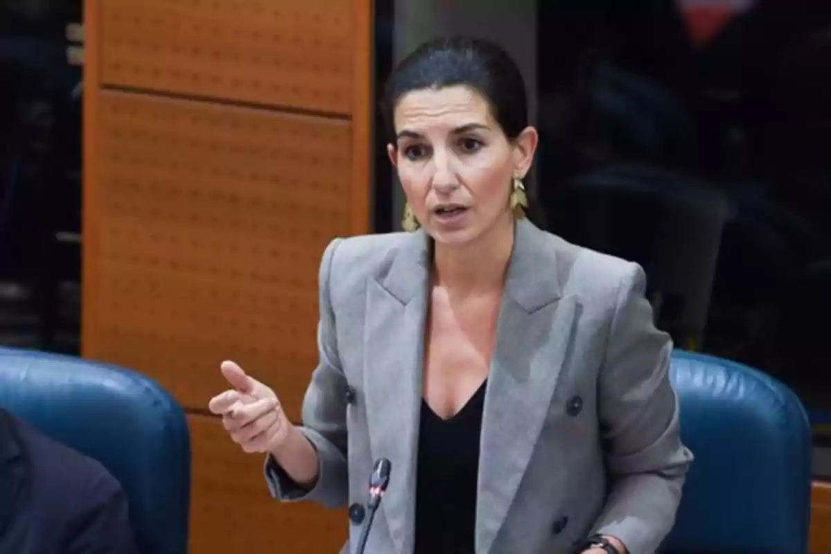 Mujer de cabello oscuro recogido, vestida con saco gris, hablando y gesticulando en lo que parece ser una sala de reuniones o parlamento Mujer de cabello oscuro recogido, vestida con saco gris, hablando y gesticulando en lo que parece ser una sala de reuniones o parlamento