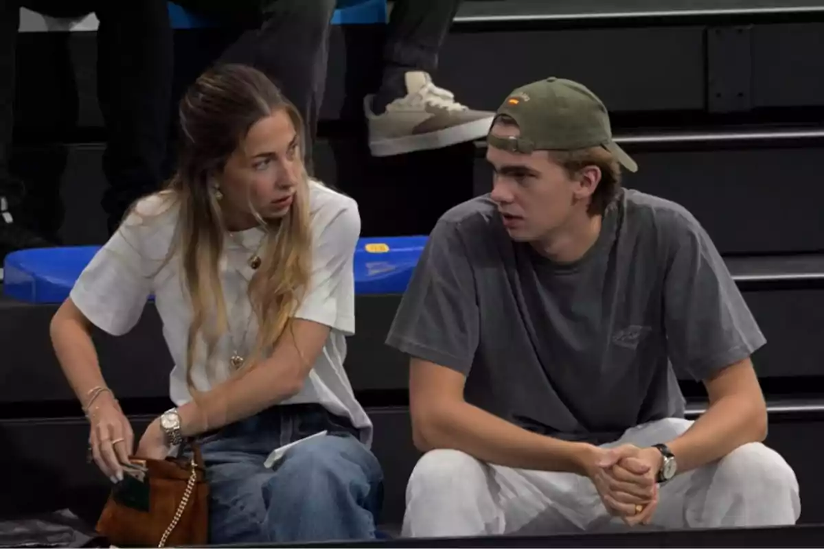 Dos personas sentadas en unas gradas conversando, una mujer con camiseta blanca y bolso marrón y un hombre con gorra verde y camiseta gris. Dos personas sentadas en unas gradas conversando, una mujer con camiseta blanca y bolso marrón y un hombre con gorra verde y camiseta gris.