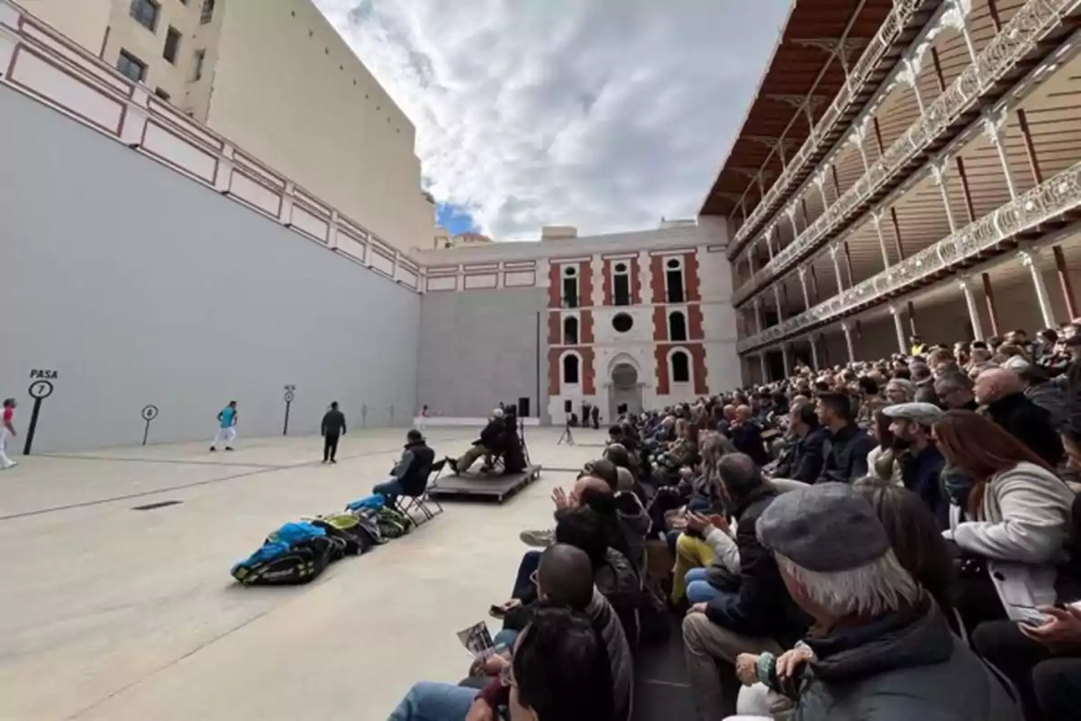Personas observan un partido de pelota vasca en un frontón rodeado de gradas y una fachada histórica