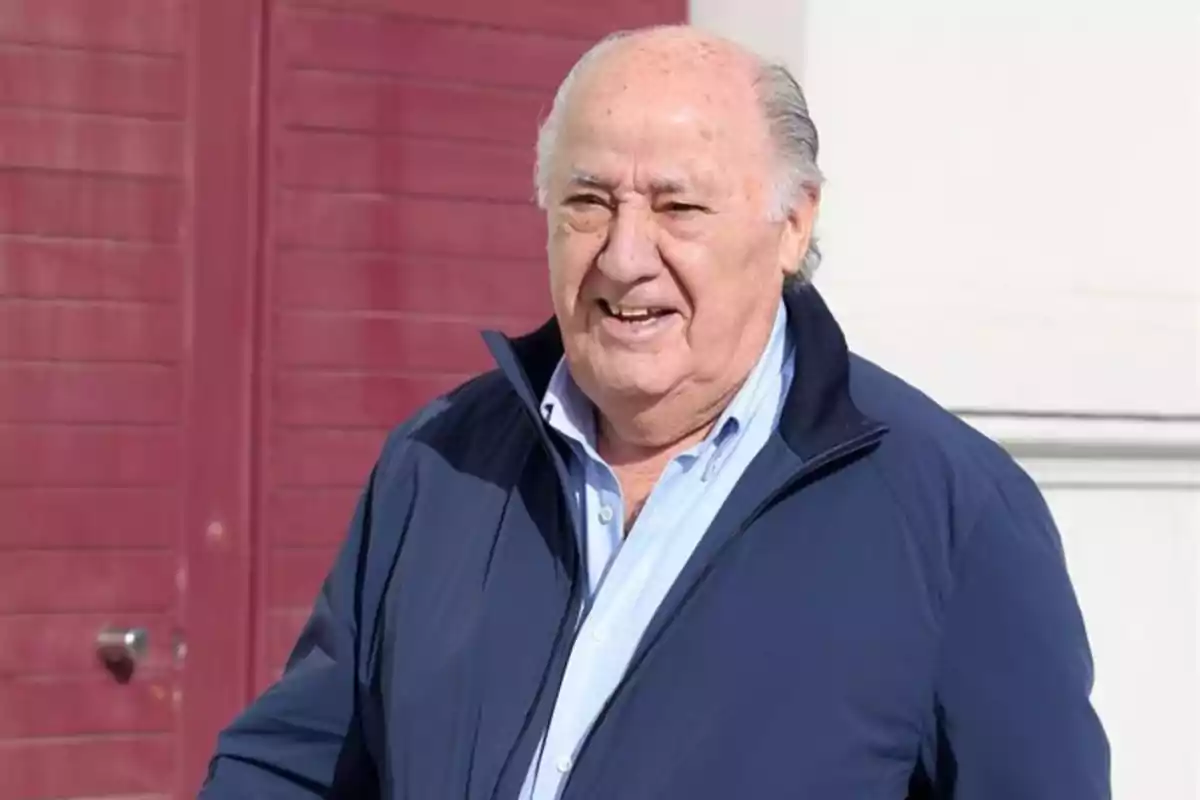 Un hombre mayor con chaqueta azul y camisa clara sonriendo frente a una puerta roja.