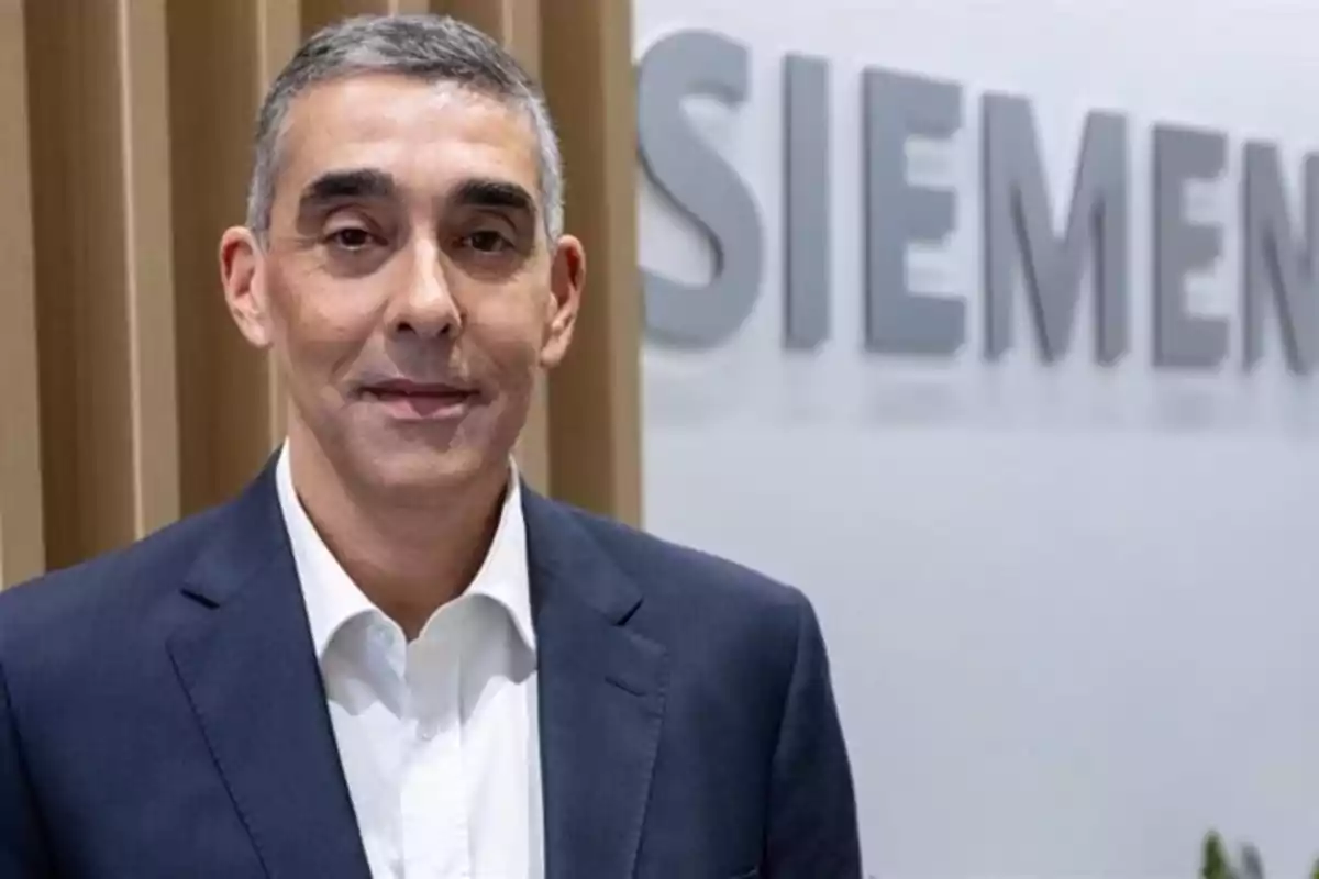 Hombre de cabello corto y canoso con traje azul y camisa blanca posando frente a un letrero de Siemens Hombre de cabello corto y canoso con traje azul y camisa blanca posando frente a un letrero de Siemens