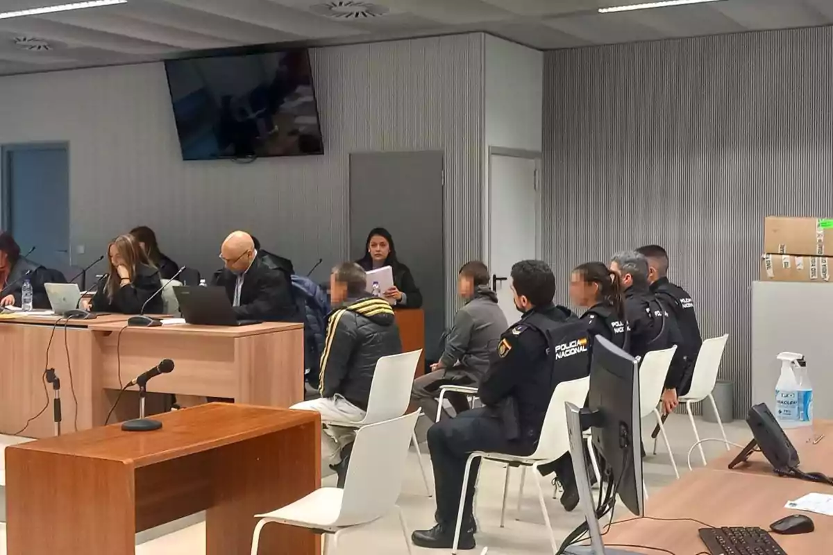 Un grupo de personas, incluyendo oficiales de policía, se encuentra en una sala de reuniones o tribunal, sentados alrededor de mesas con computadoras y documentos.