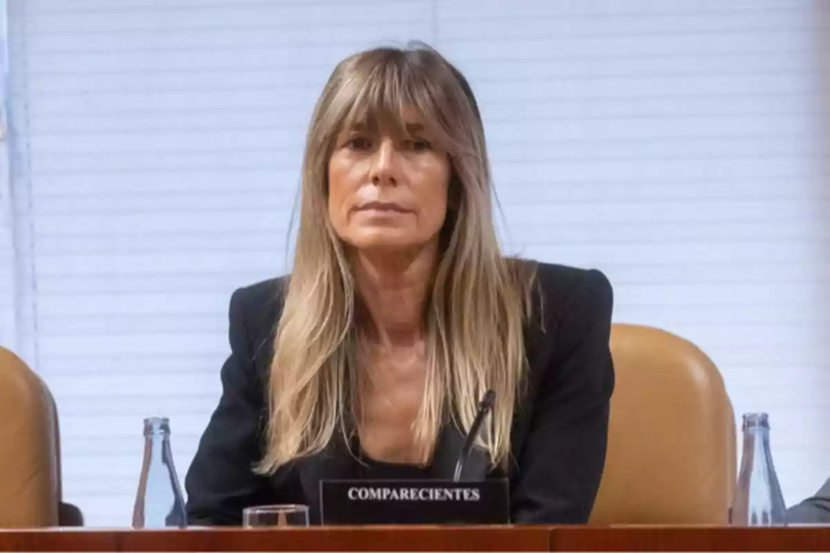 Una mujer con cabello rubio y flequillo está sentada en una sala de conferencias con un cartel que dice "COMPARECIENTES" frente a ella.