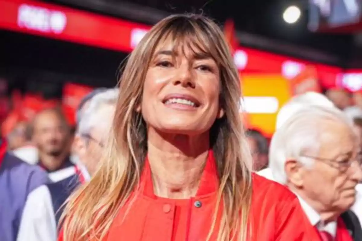 Una mujer sonriente con chaqueta roja en un evento con luces y personas de fondo.