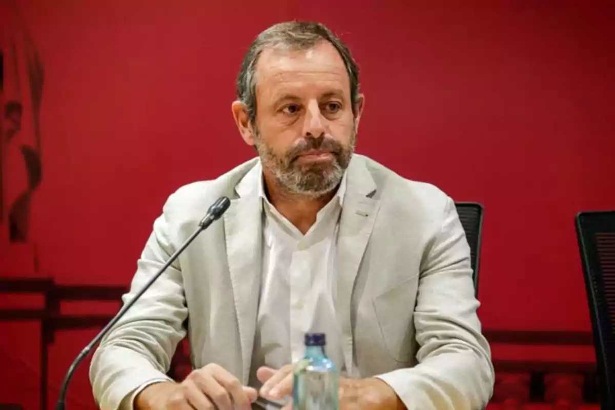 Hombre con barba y cabello corto, vestido con traje claro, sentado frente a un micrófono sobre un fondo rojo.