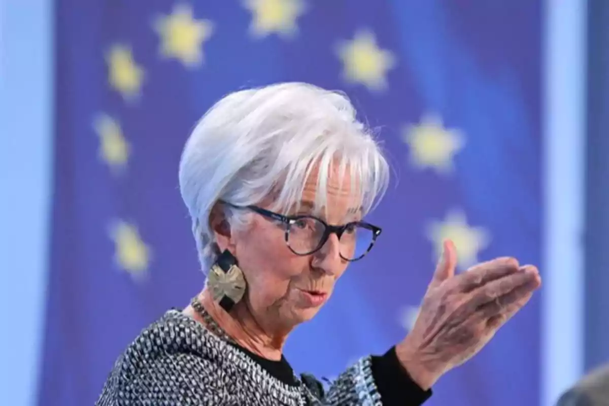 Una persona de cabello blanco y gafas gesticula con la mano frente a un fondo con estrellas amarillas sobre un fondo azul.