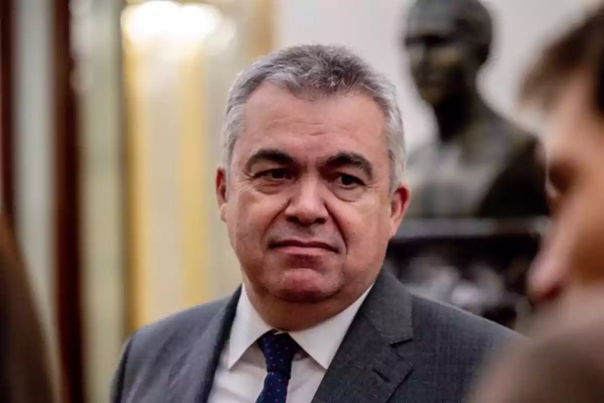 Hombre de cabello canoso con traje y corbata en un entorno formal con una escultura al fondo Hombre de cabello canoso con traje y corbata en un entorno formal con una escultura al fondo