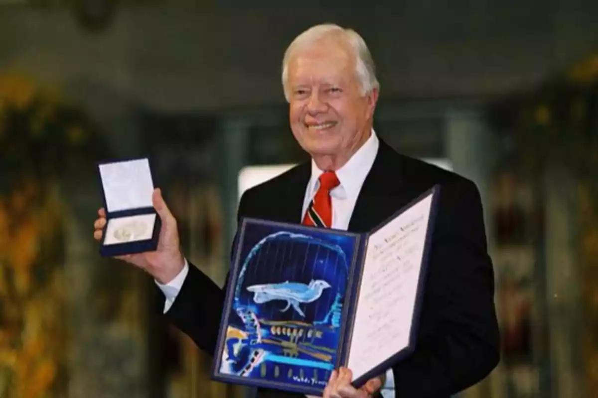 Un hombre mayor sonriente sostiene un diploma y una medalla en un estuche abierto.