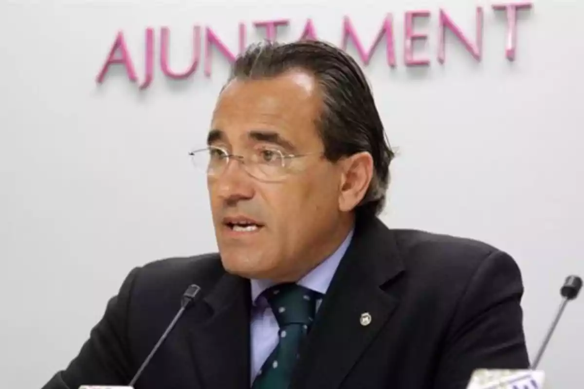 Un hombre con gafas y traje oscuro hablando frente a un micrófono con la palabra "AJUNTAMENT" en el fondo.