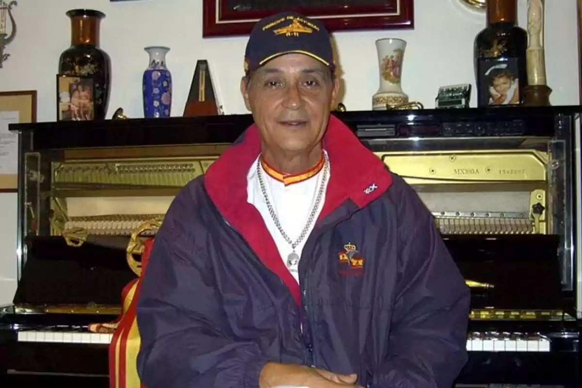 Un hombre con gorra y chaqueta está sentado frente a un piano, rodeado de varios objetos decorativos.