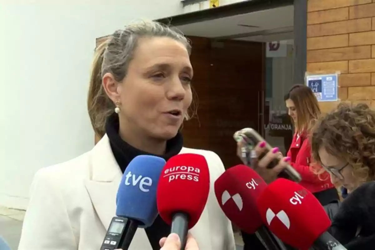 Una mujer hablando frente a varios micrófonos de prensa en un entorno exterior. Una mujer hablando frente a varios micrófonos de prensa en un entorno exterior.