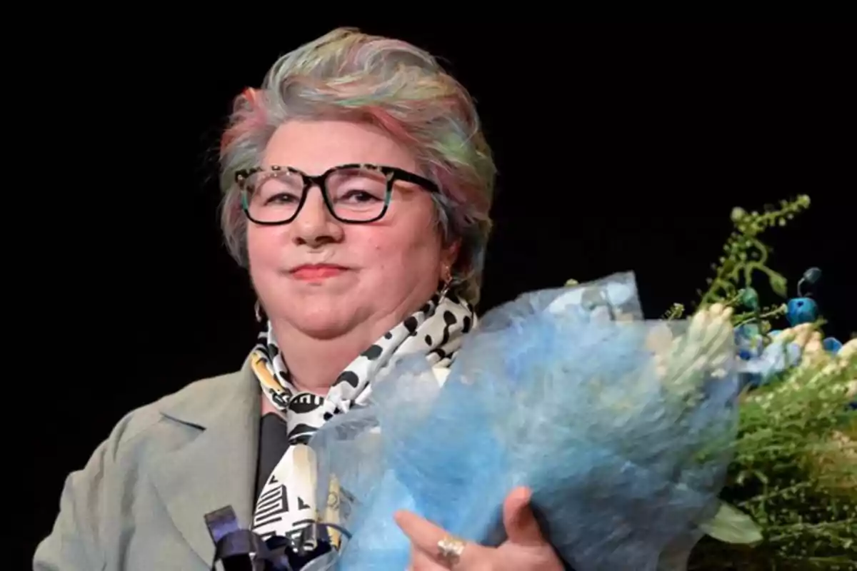 Una persona con cabello de colores y gafas sostiene un ramo de flores azules. Una persona con cabello de colores y gafas sostiene un ramo de flores azules.