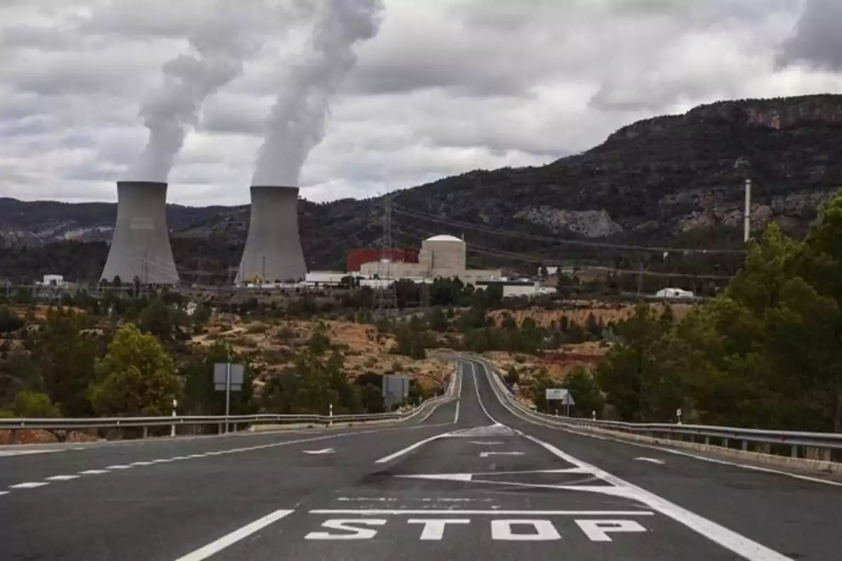 Carretera con señal de stop en primer plano y al fondo una planta industrial con dos torres de refrigeración emitiendo vapor, rodeada de montañas y vegetación. Carretera con señal de stop en primer plano y al fondo una planta industrial con dos torres de refrigeración emitiendo vapor, rodeada de montañas y vegetación.