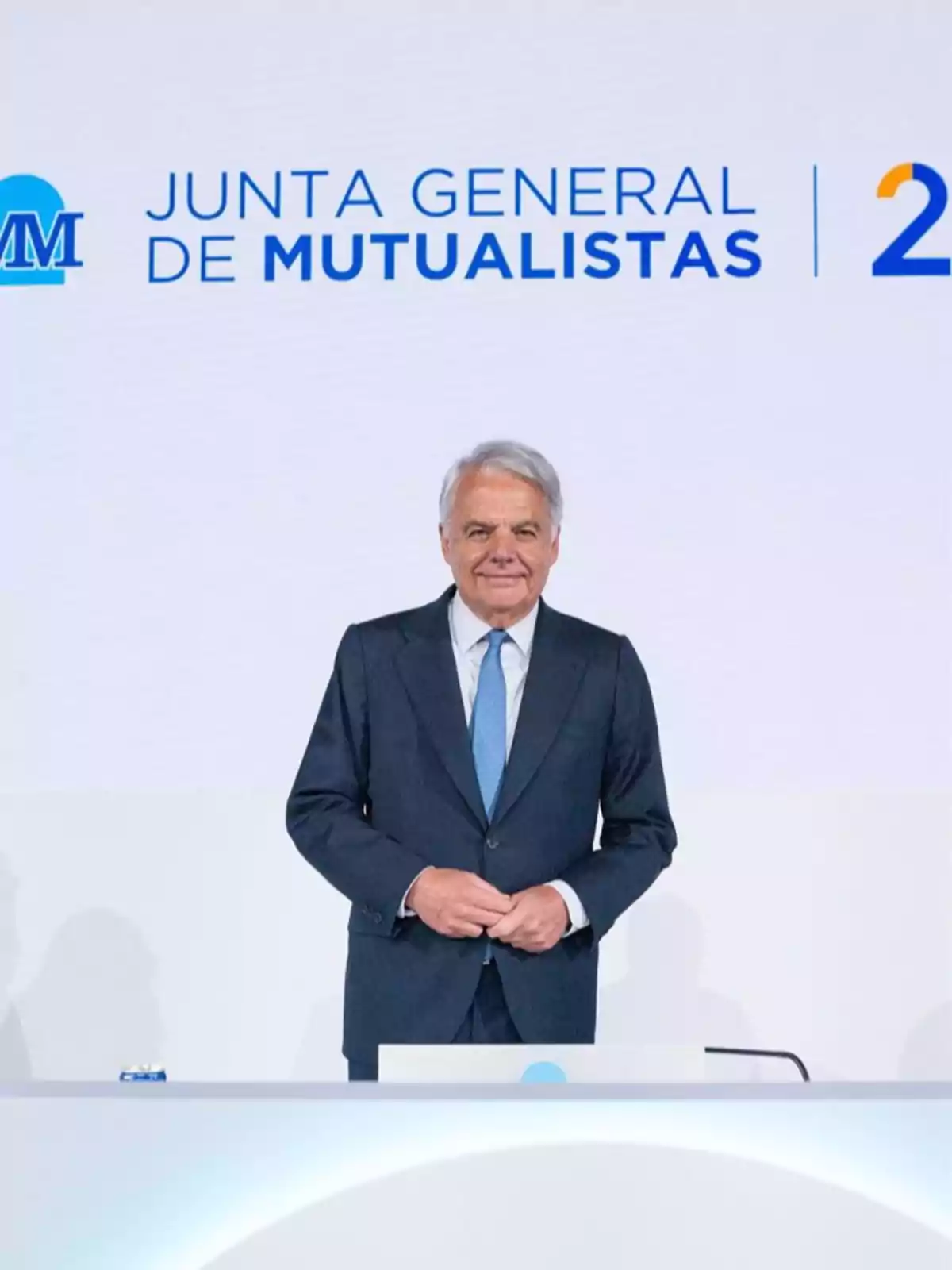 Hombre de cabello canoso con traje oscuro y corbata azul de pie frente a un podio en un evento de la Junta General de Mutualistas Hombre de cabello canoso con traje oscuro y corbata azul de pie frente a un podio en un evento de la Junta General de Mutualistas
