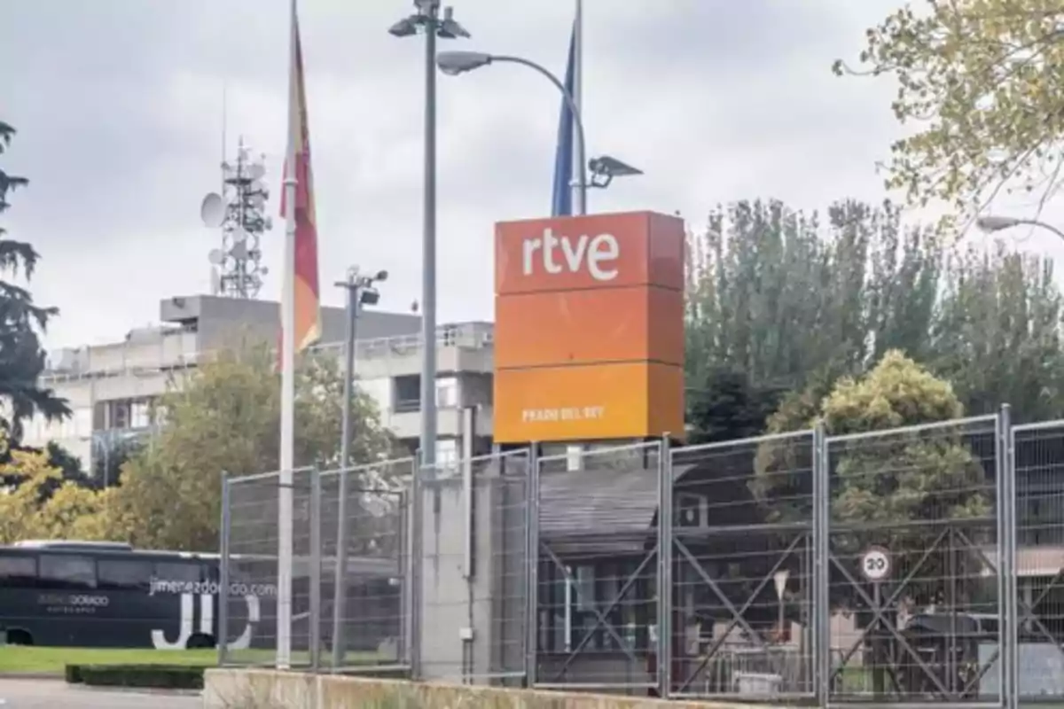 Entrada principal de un edificio de RTVE con un cartel naranja y banderas al frente. Entrada principal de un edificio de RTVE con un cartel naranja y banderas al frente.