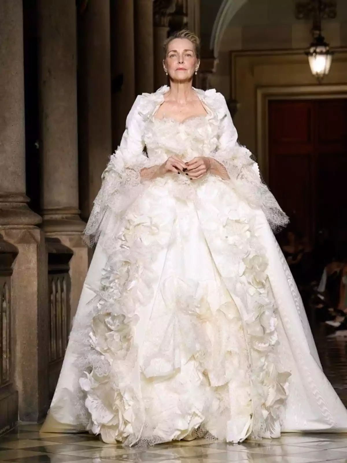 Mujer elegante desfilando con un vestido blanco voluminoso decorado con flores y encaje en una pasarela de interiores Mujer elegante desfilando con un vestido blanco voluminoso decorado con flores y encaje en una pasarela de interiores