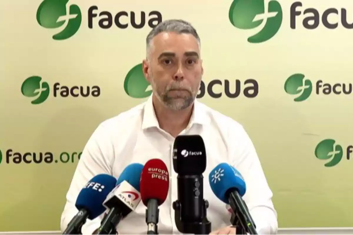 Un hombre con camisa blanca está sentado frente a varios micrófonos de diferentes medios de comunicación con un fondo que tiene el logo de Facua. Un hombre con camisa blanca está sentado frente a varios micrófonos de diferentes medios de comunicación con un fondo que tiene el logo de Facua.