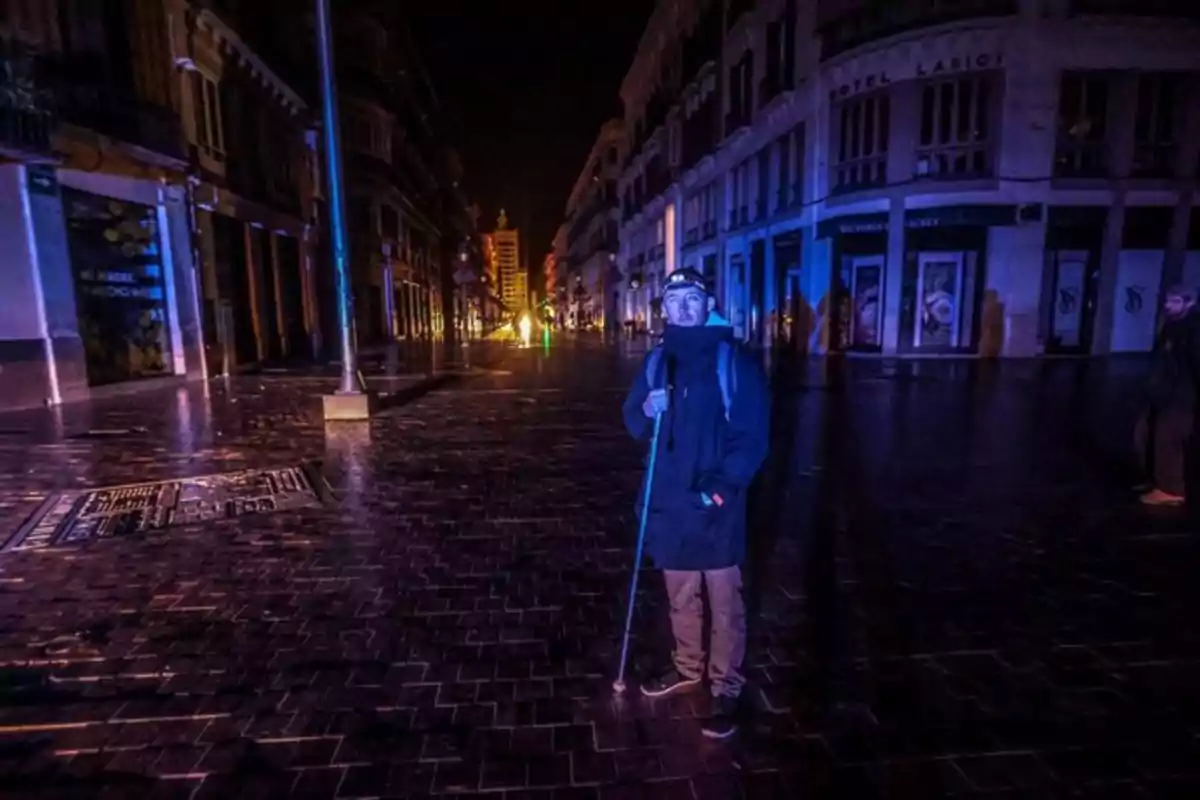 Un hombre con una linterna en la cabeza y un bastón está de pie en una calle oscura y vacía, rodeado de edificios iluminados tenuemente. Un hombre con una linterna en la cabeza y un bastón está de pie en una calle oscura y vacía, rodeado de edificios iluminados tenuemente.