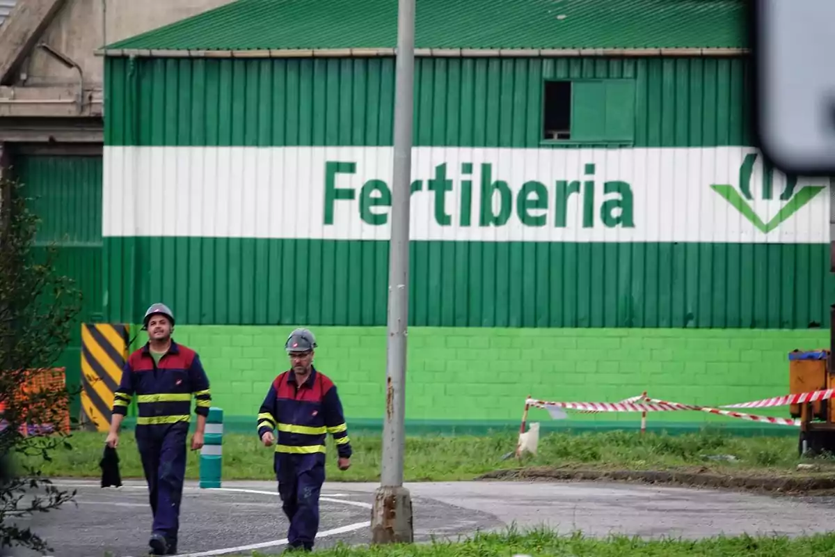 Dos trabajadores con cascos y uniformes de seguridad caminan frente a un edificio verde con el letrero "Fertiberia".