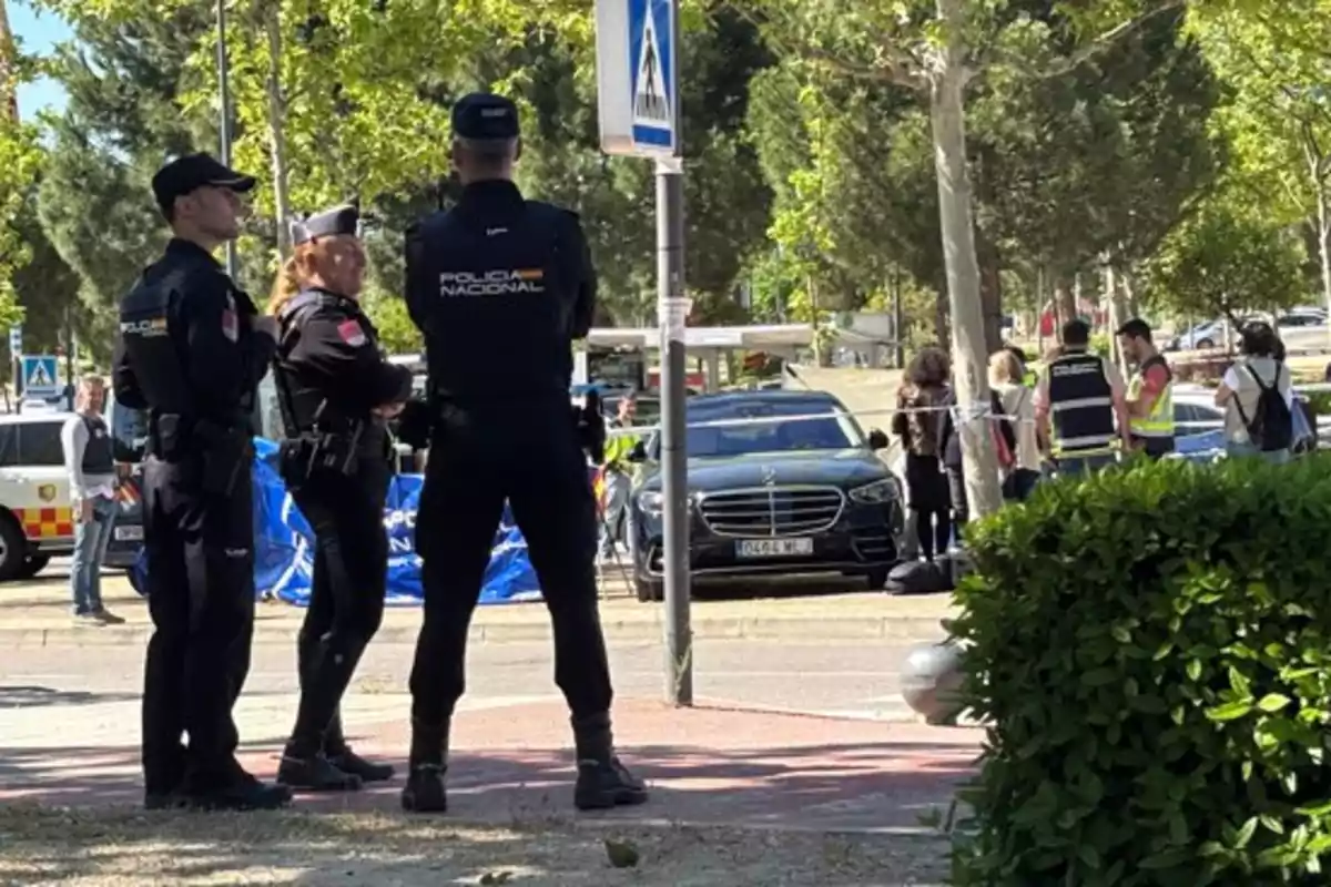 Un grupo de policías nacionales está de pie en una calle acordonada mientras varias personas observan y un coche negro está estacionado cerca. Un grupo de policías nacionales está de pie en una calle acordonada mientras varias personas observan y un coche negro está estacionado cerca.