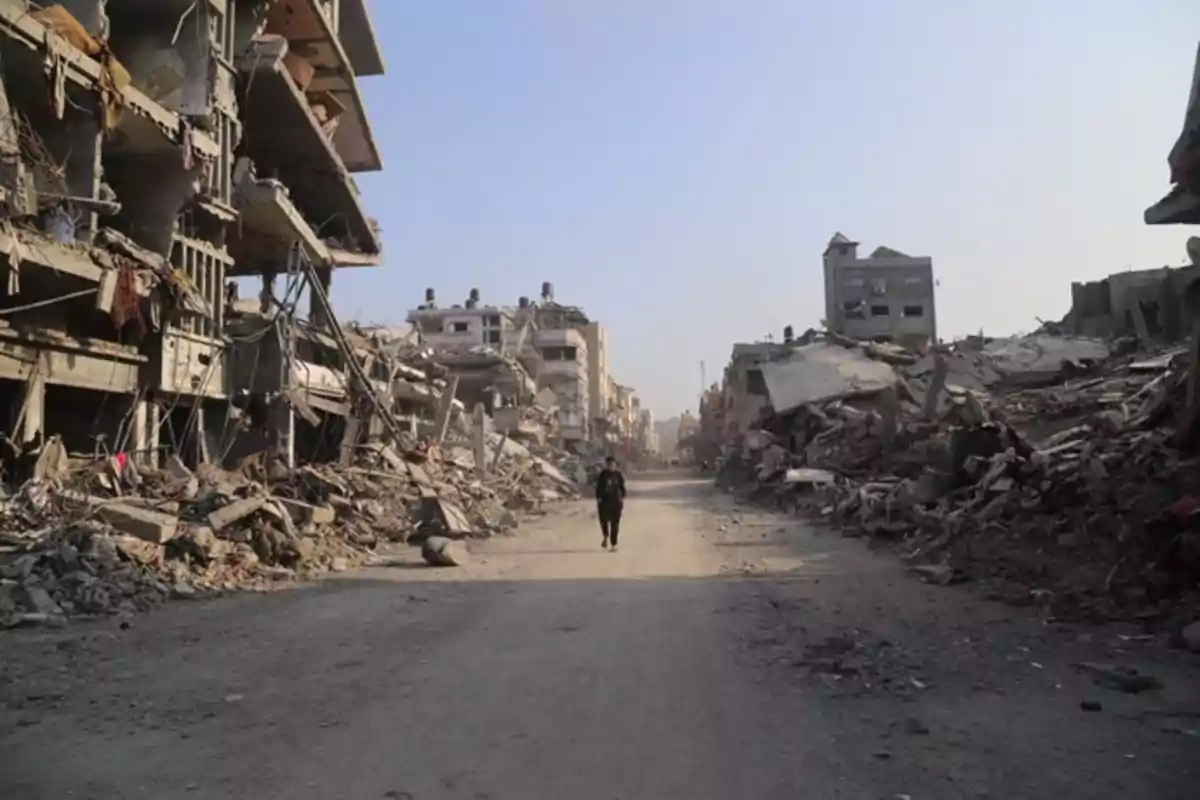 Una persona camina por una calle rodeada de edificios en ruinas y escombros bajo un cielo despejado. Una persona camina por una calle rodeada de edificios en ruinas y escombros bajo un cielo despejado.